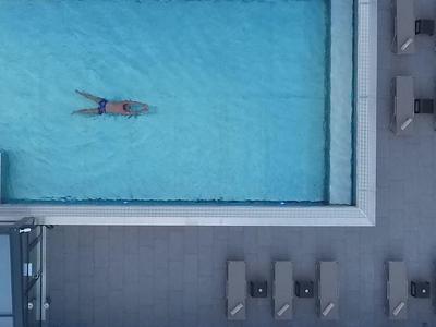 A person floats in a pool beside a deck with lounge chairs and shaded seating.