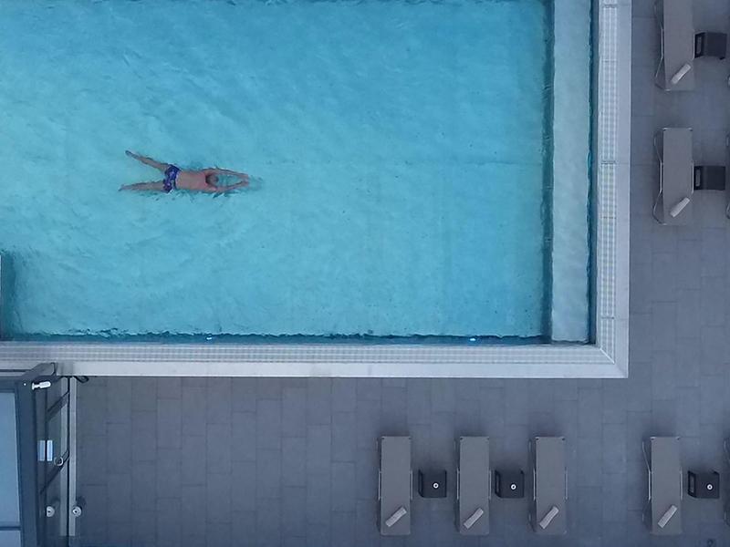 A person floats in a pool beside a deck with lounge chairs and shaded seating.
