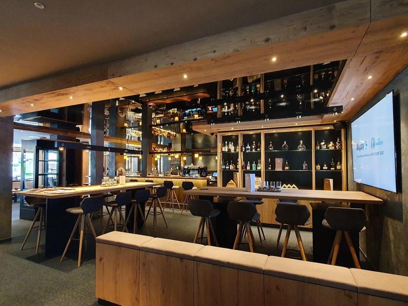Modern hotel bar with wooden ceiling, shelves of bottles, and sleek black stools.