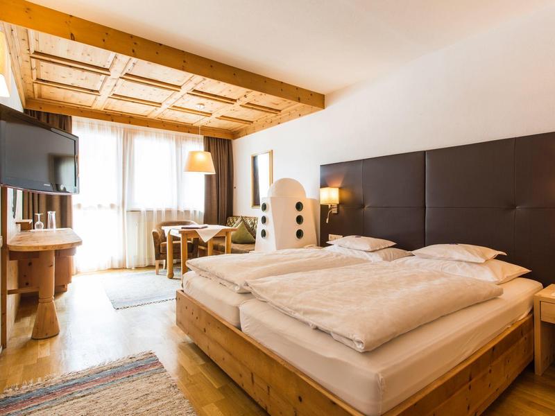 Bright hotel room with wooden furniture, a large bed, TV, and desk by the window.