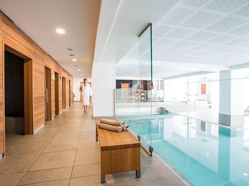 Indoor hotel pool area with wooden walls, tiled floor, bench, and a woman walking in the background.