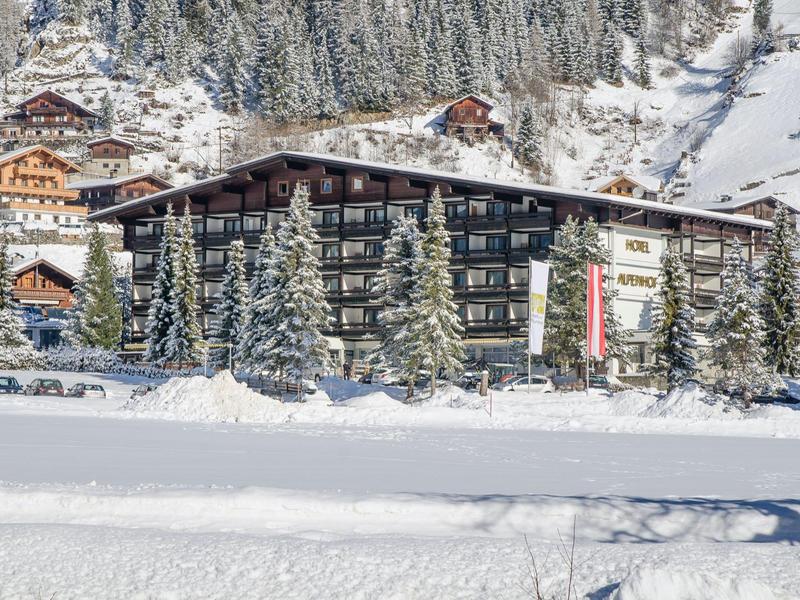 Großes Hotelgebäude mit Holzbalkonen, schneebedeckte Bäume und Winterlandschaft im Hintergrund.