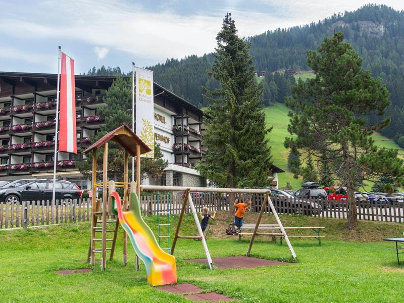 Kinderspielplatz mit Rutsche, Schaukel und Tisch vor Bergen und Hotelgebäude im Hintergrund.