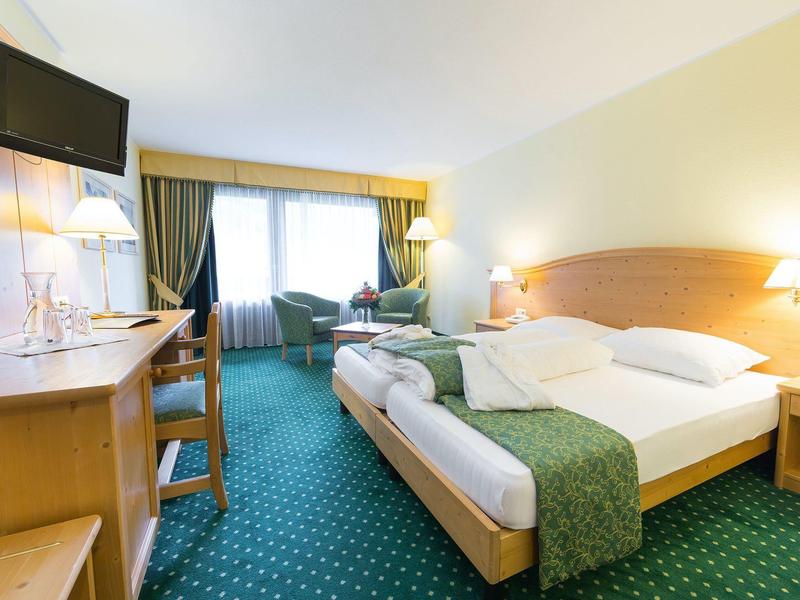 Hell eingerichtetes Hotelzimmer mit grünem Teppich, Doppelbett, Schreibtisch und Fenster mit Vorhängen.