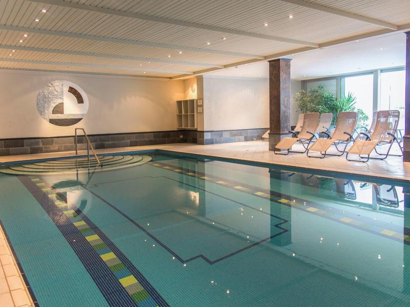 Moderner Hallenbadbereich mit klarem Wasser, Liegestühlen und großen Fenstern für Tageslicht.