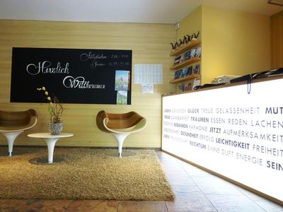 Moderner Wartebereich mit weißen Stühlen, Holzpaneel, Teppich und beleuchtetem Zitat an der Wand.