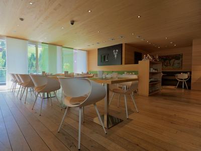 Ristorante moderno in hotel con pavimento in legno, sedie bianche e grandi finestre con vista sul verde.