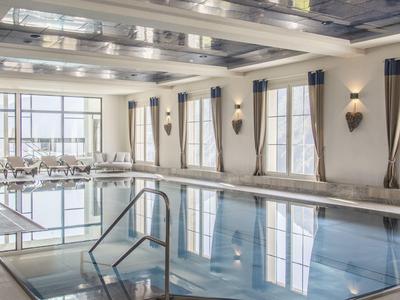 Heller Innenpool mit großen Fenstern, grauen Vorhängen und weißen Liegestühlen am Rand.