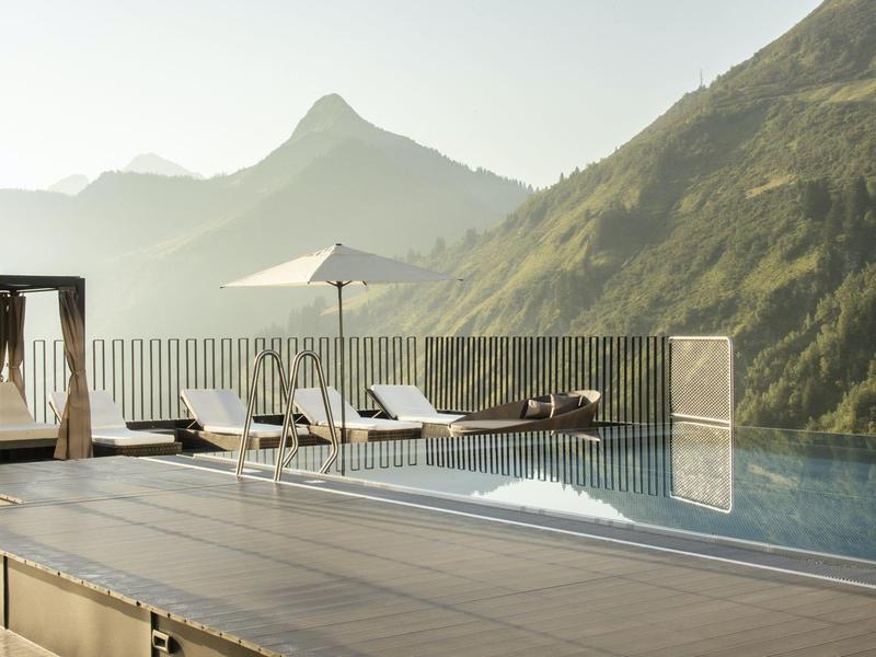 Infinity-Pool mit Liegestühlen und Sonnenschirm, umgeben von Bergen im sanften Morgenlicht.
