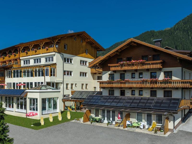 Velký hotel s bílými a dřevěnými budovami před zalesněnými horami za jasné oblohy.