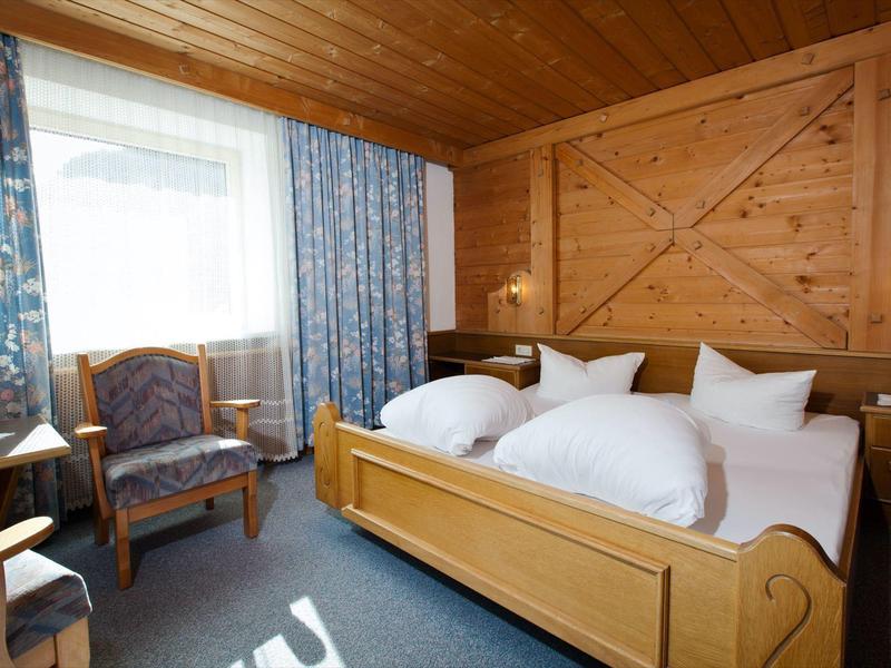 Jasny pokój hotelowy z drewnianym sufitem, podwójnym łóżkiem, dwoma fotelami i niebieskimi zasłonami w wzory.