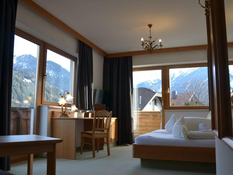 Habitación de hotel luminosa con grandes ventanas, muebles de madera, cama y escritorio, vista a las montañas.