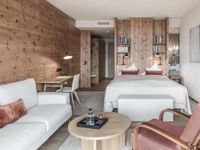 Modern hotel room with bed, sofa, armchair, and wooden accents in warm tones.