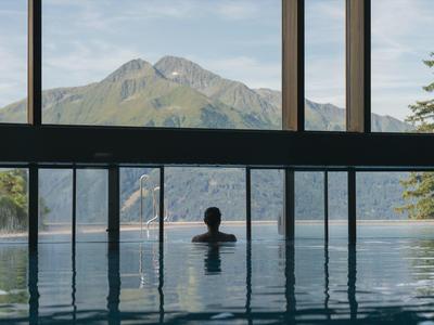 Person in an infinity pool overlooking mountains and lake under a cloudy sky.