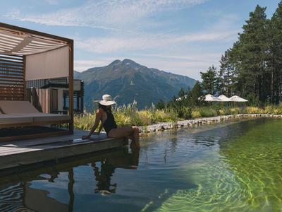 Vrouw zit op een aanlegsteiger naast een meer met bergachtergrond en een modern open paviljoen.