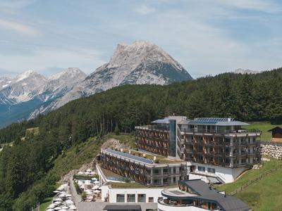 Modern hotel with terrace and lounge chairs in front of forested mountains and peaks.