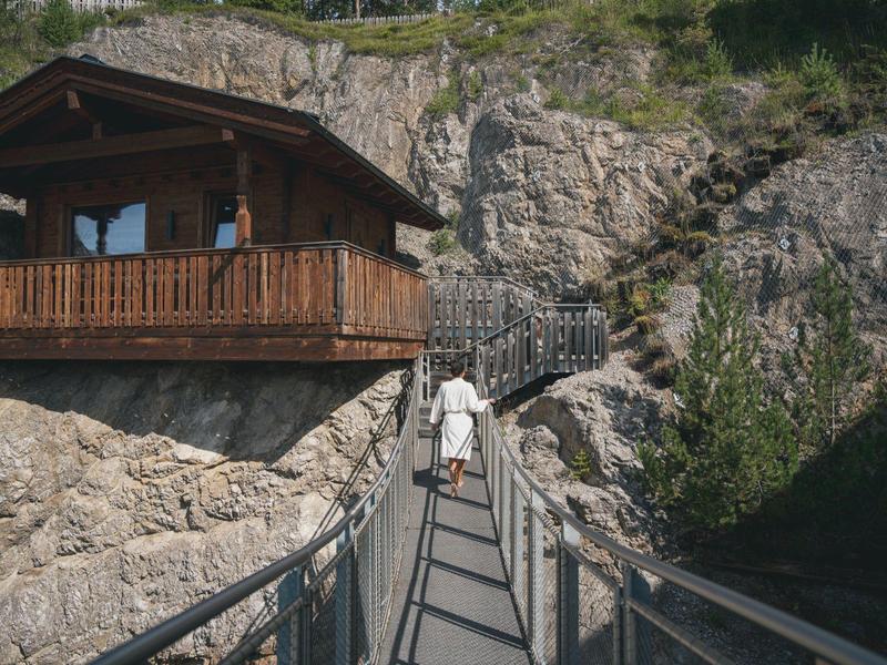 Person walks over a bridge to a wooden cabin nestled in rock formations.