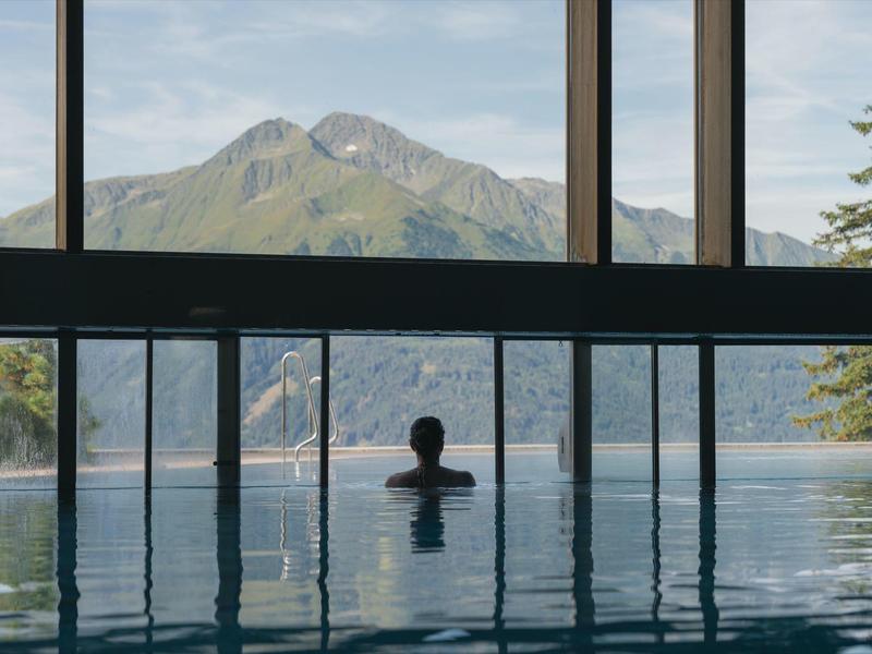 Person in an infinity pool overlooking mountains and lake under a cloudy sky.