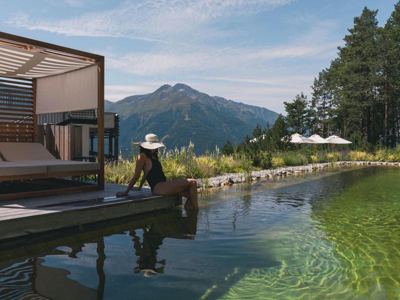 Person sits on a dock by a clear pond with mountains and trees in the background.