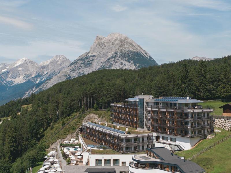 Modern hotel with terrace and lounge chairs in front of forested mountains and peaks.