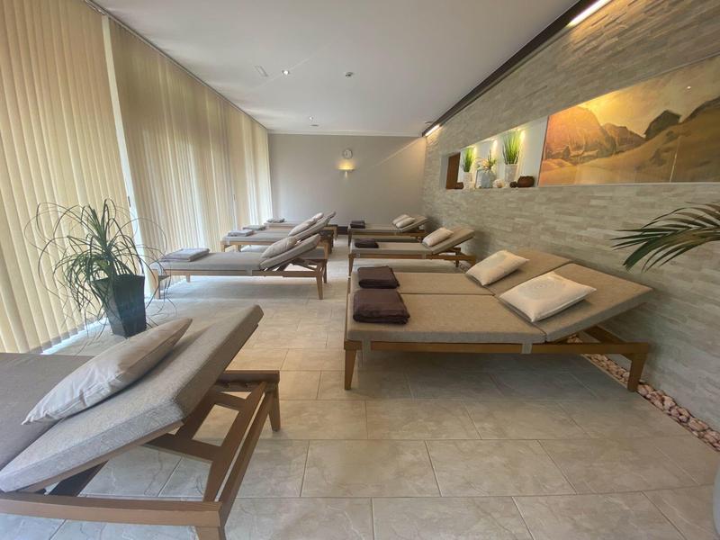 Sala relax luminosa con lettini e grandi finestre, decorata con piante e quadri.