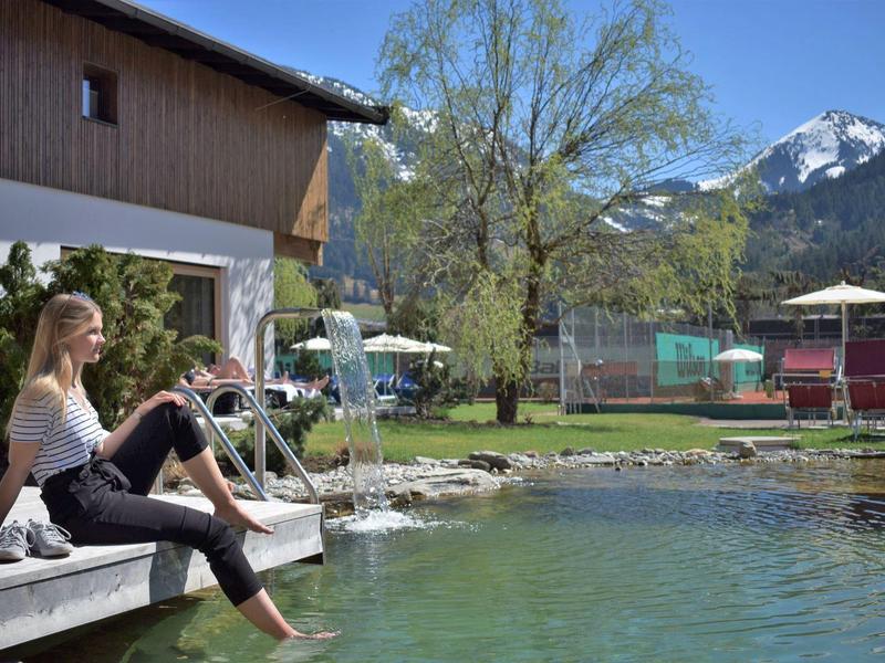 Frau sitzt entspannt am Teichufer vor einem Holzhaus mit Bergblick und grünem Garten.