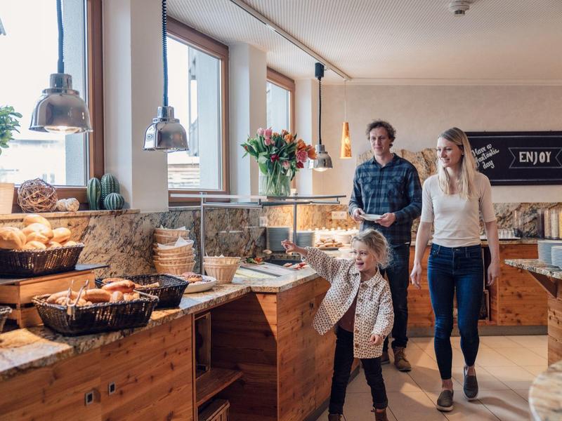 Familie läuft in helles Café mit Holztischen, Brotkörben und großen Fenstern voller Tageslicht.