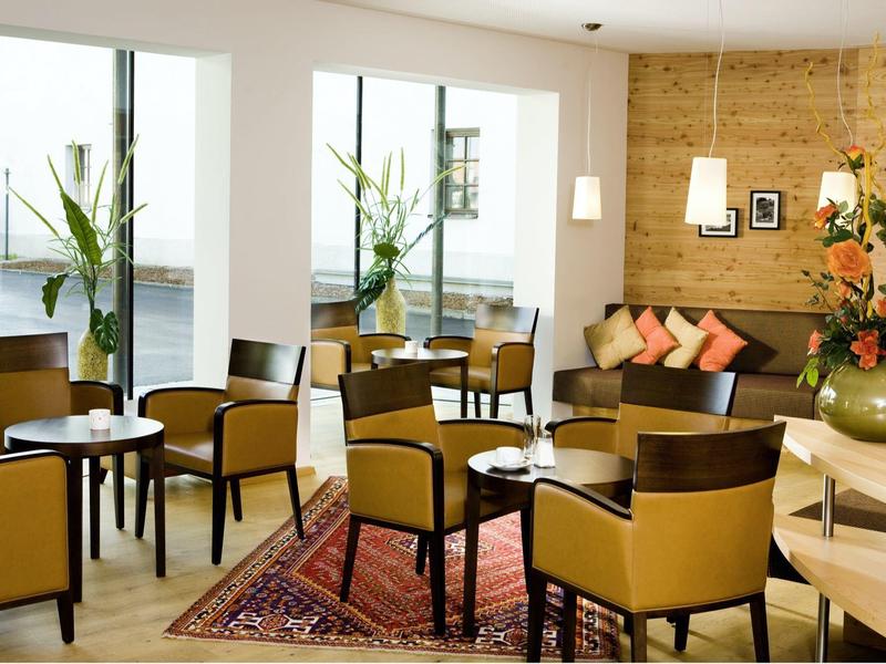 Modernes Café mit beigen Sesseln, Holzlattenwand, bunten Kissen und großen Pflanzenarrangements.