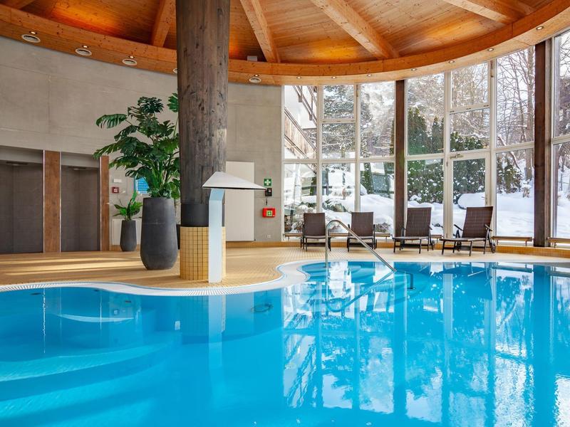 Innenraum mit rundem Schwimmbecken, Holzdecke, großen Fenstern und Entspannungsbereich mit Stühlen.