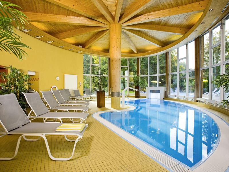 Innenliegendes Schwimmbad mit gebogenem Becken, Holzdecke und Liegestühlen neben großen Fenstern.