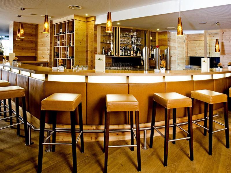 Moderne Bar mit hölzernen Hockern, warmem Licht und hellen Holz- und Steinwänden.
