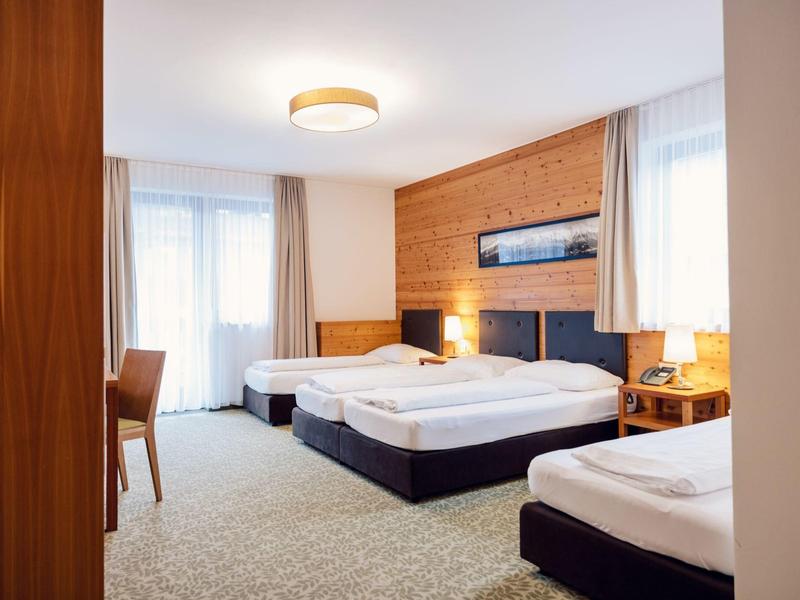 Helles Hotelzimmer mit drei Einzelbetten, Holzdekor und großem Fenster mit Vorhängen.