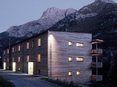 Modernes Holzhaus mit Beleuchtung vor dunklem Bergpanorama bei Dämmerung.
