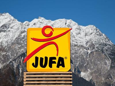 Logo JUFA jaune devant des montagnes enneigées et un ciel bleu clair.