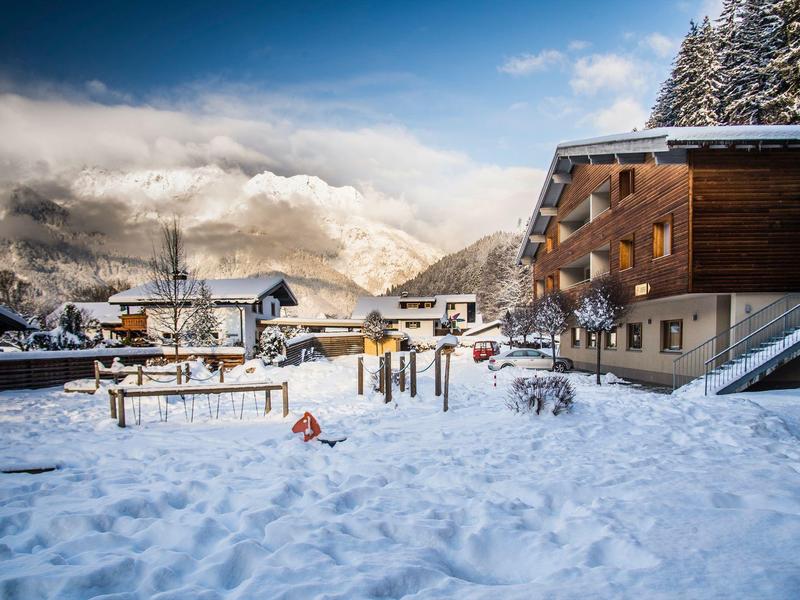 Snow-covered mountain village with wooden chalets and playground under cloudy sky.