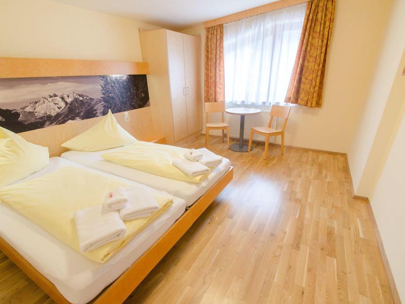 Bright hotel room with double bed, wooden floor, two chairs, and window with curtains.