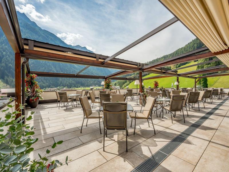 Terrace with tables and chairs overlooking mountains, sunny with open glass roof.