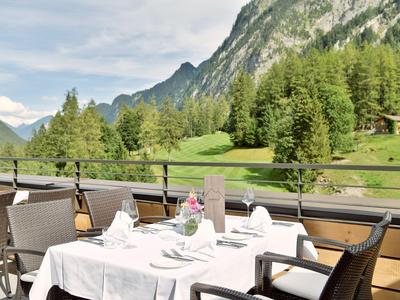 Terrasse avec tables dressées offrant vue sur prairies verdoyantes et montagnes sous un ciel clair.