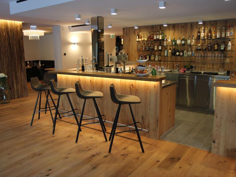 Modern hotel bar area with wooden counter, illuminated seats, and many bottles in the background.