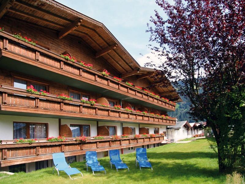 Maison en bois à plusieurs étages avec balcons et chaises longues bleues sur pelouse verte sous ciel bleu.