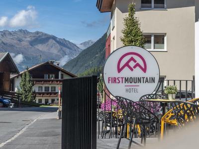 Straßenszene mit einem Schild des First Mountain Hotels vor Bergkulisse bei klarem Himmel