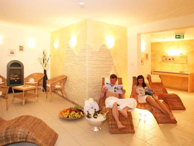 Zwei Personen entspannen auf Liegestühlen in einem hellen Wellnessraum mit Handtüchern und Obst.