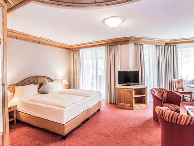 Helles Hotelzimmer mit Doppelbett, Sitzbereich, Fernseher und großen Fenstern mit Vorhängen.