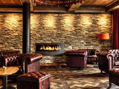 Przytulny hotelowy salon z kamienną ścianą, otwartym kominkiem i skórzanymi fotelami w ciepłym świetle.