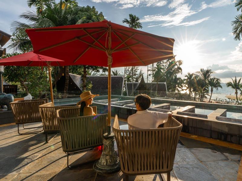 Zwei Personen sitzen auf Stühlen unter roten Sonnenschirmen nahe einem Pool mit Palmen im Sonnenuntergang.