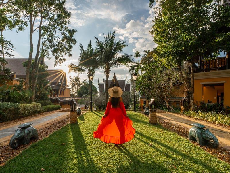Frau im roten Kleid geht auf einem grünen Weg, umgeben von Bäumen und Gebäuden bei Sonnenuntergang.