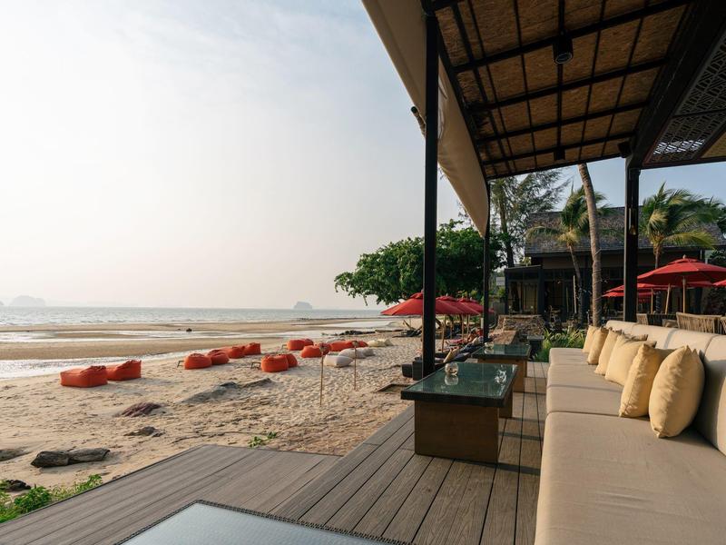 Strand mit gemütlicher Terrasse, bequemen Kissen und roten Sonnenschirmen bei ruhigem Meer.