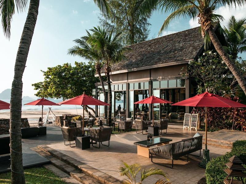Restaurantterrasse mit roten Sonnenschirmen, Sitzgruppen und Palmen am Meer unter blauem Himmel.