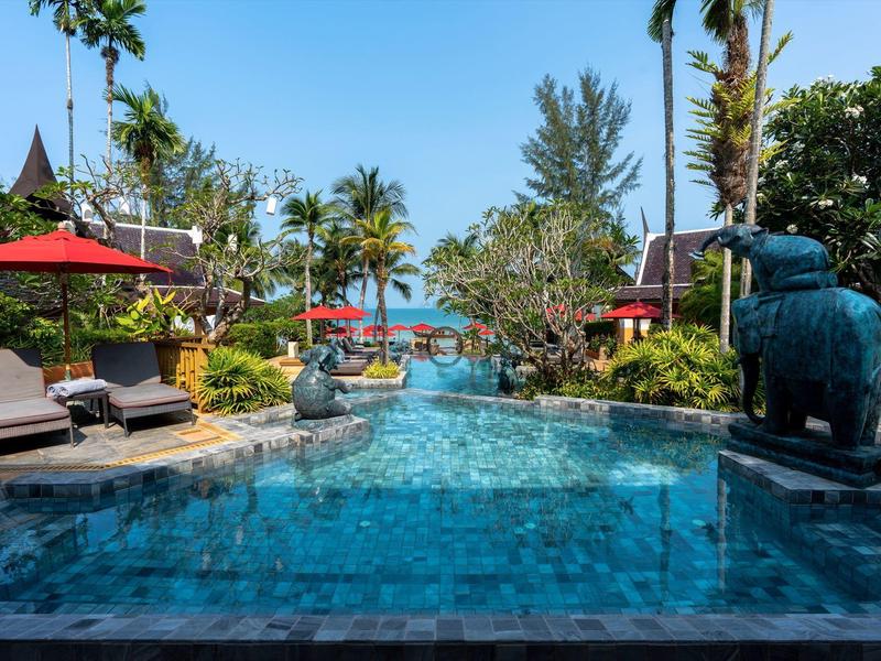 Schwimmbad mit blauem Wasser, Liegestühlen, roten Sonnenschirmen und tropischen Pflanzen im Sonnenschein.