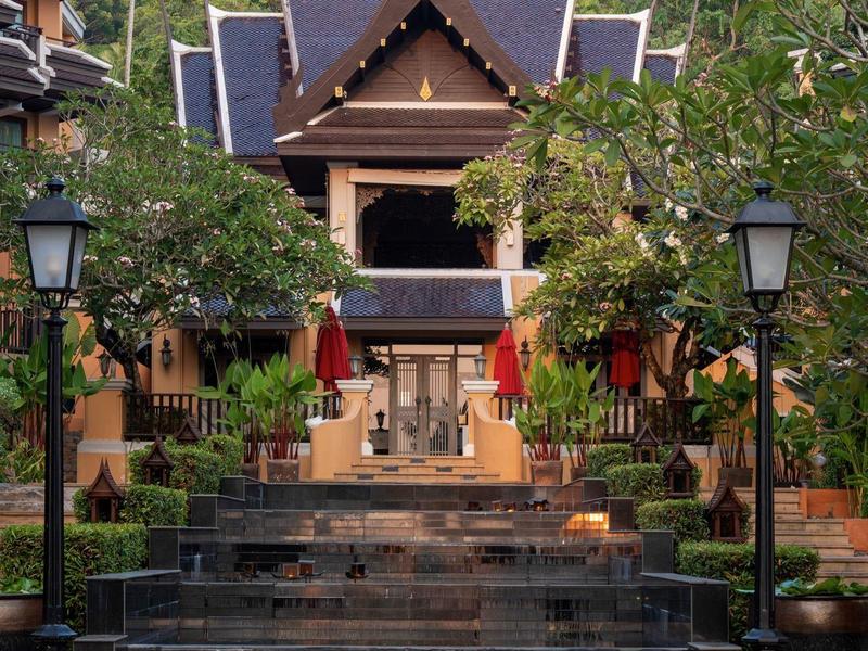 Traditionelles thailändisches Gebäude mit Treppe, Laternen und grüner Vegetation im Hintergrund.
